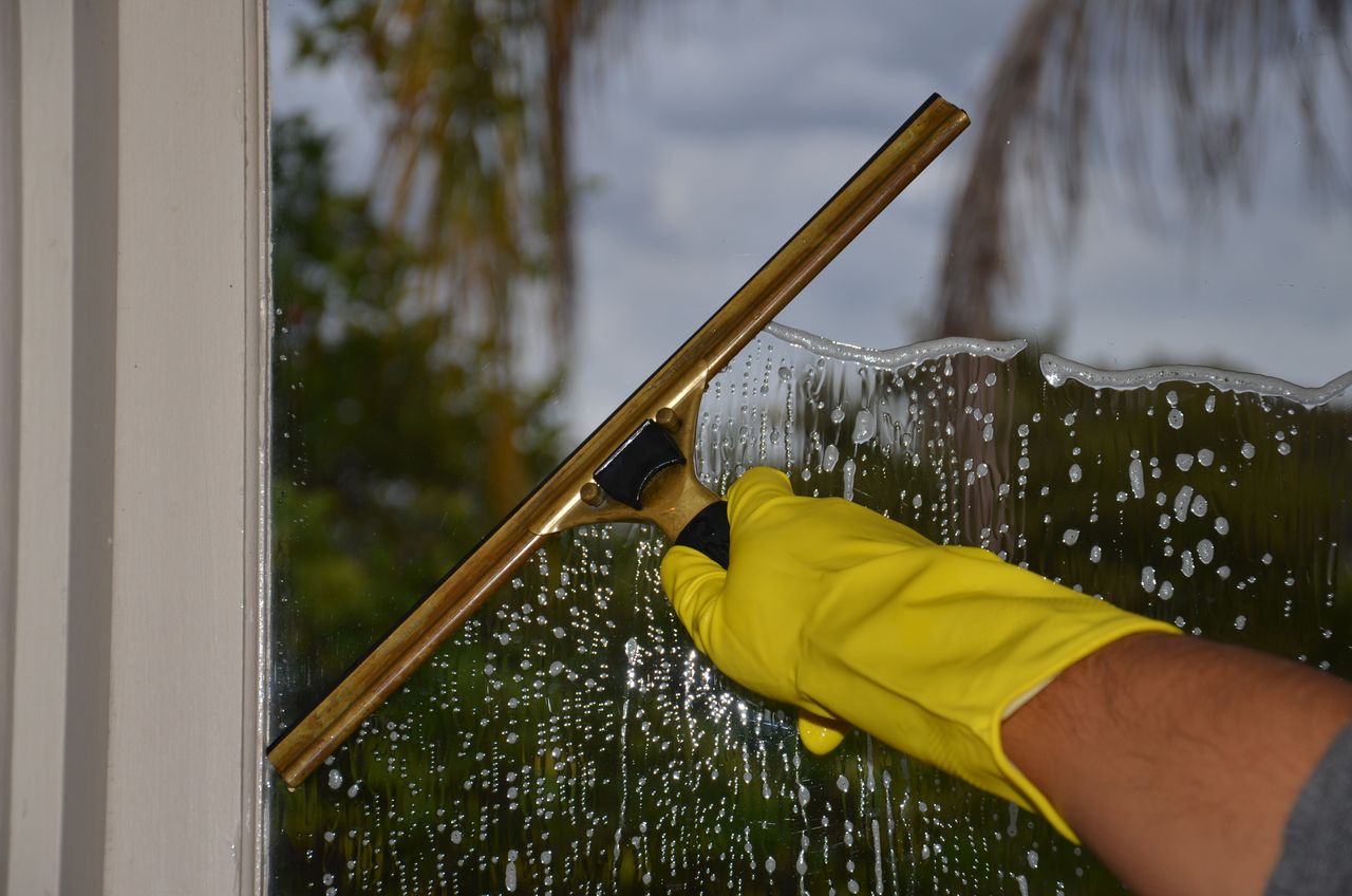 services-03 Close-up of hand cleaning window with squeegee outdoors using yellow gloves.